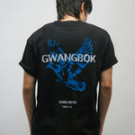 Person wearing a black t-shirt with blue dove bird graphic and white Gwangbok text on the back against a plain background