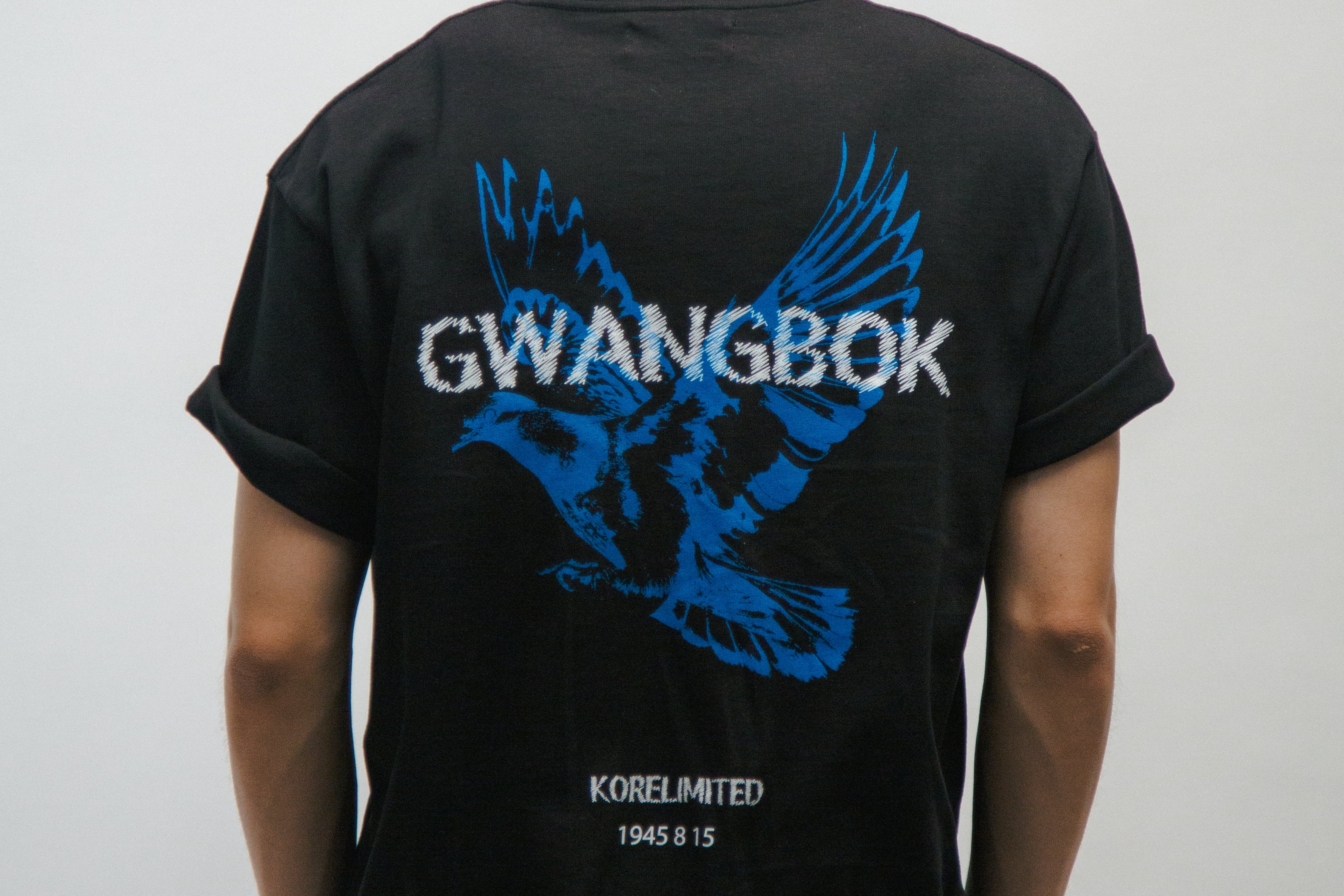 Person wearing a black t-shirt with blue dove bird graphic and white Gwangbok text on the back against a plain background