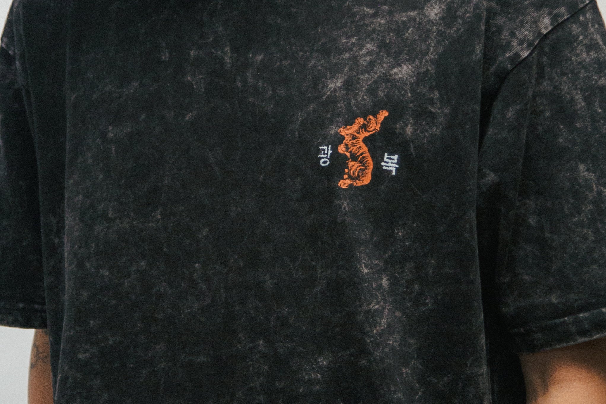 Person wearing a black acid-washed t-shirt with a small graphic of a Korean tiger and 광복 on a plain background