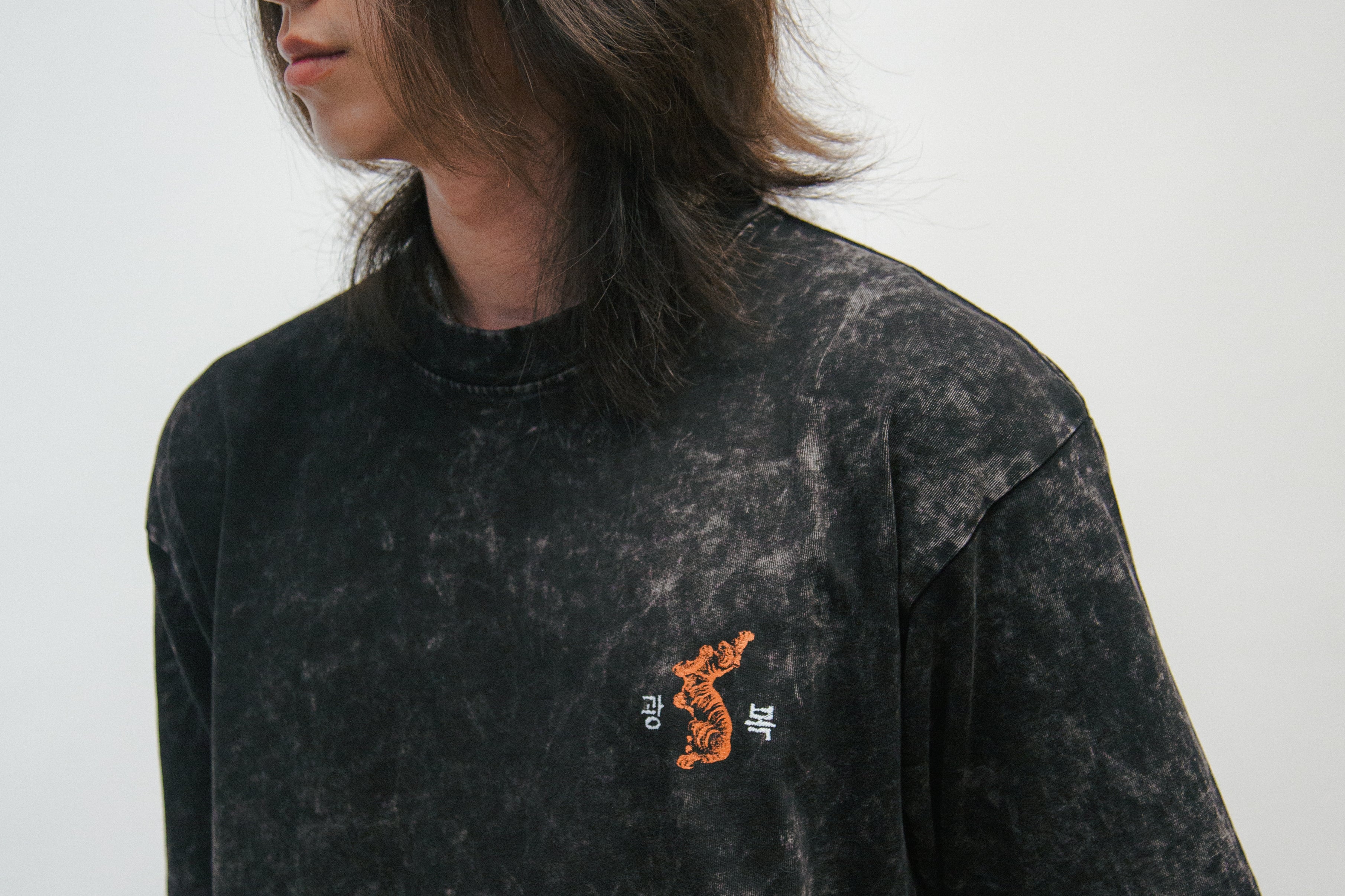 Person wearing a black acid wash t-shirt with a small graphic of an orange Korean tiger and 광복 on a plain background