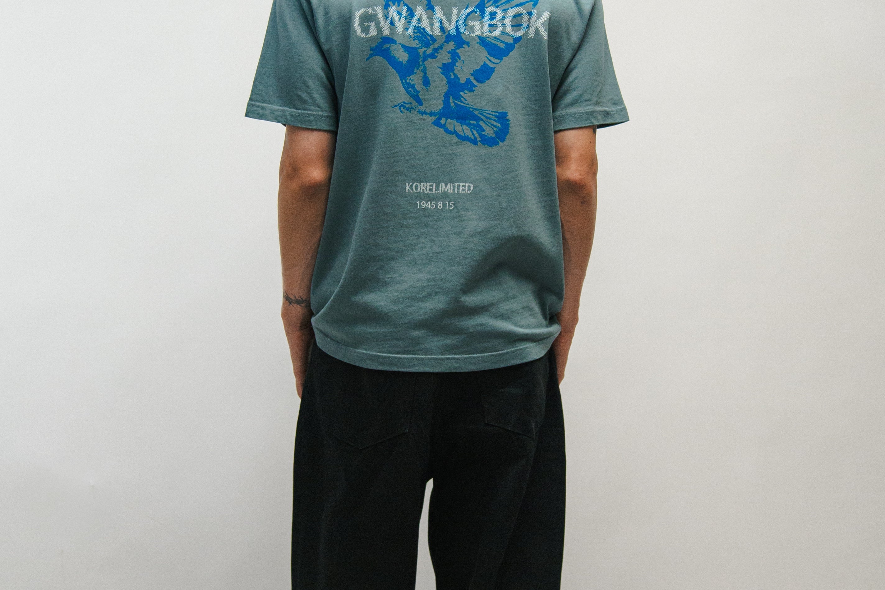 Person wearing a teal t-shirt with a blue dove logo and 'Gwangbok' on the back against a plain background