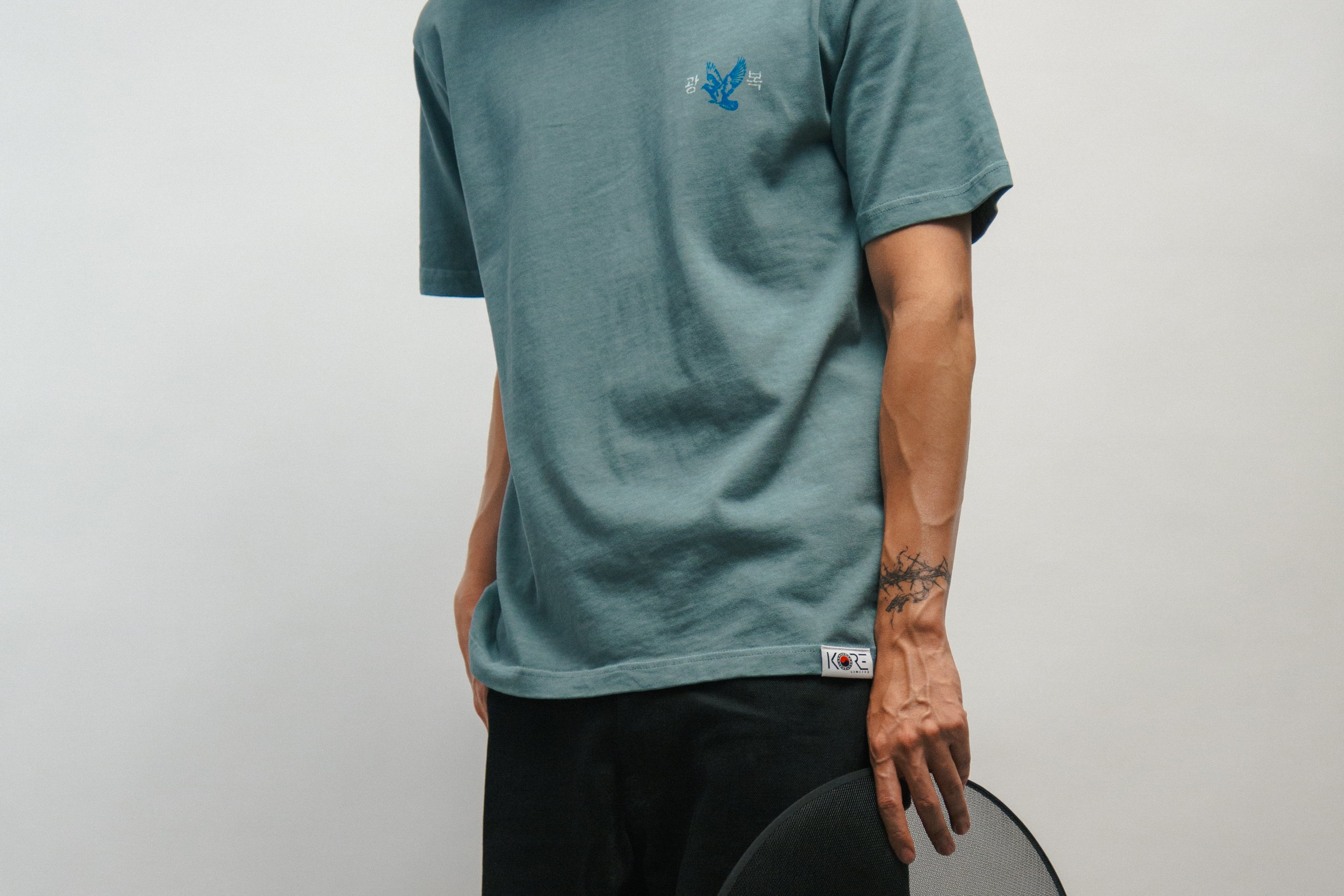 Person wearing a teal t-shirt with a logo of blue dove, holding a black Korean hat called gat against a plain background