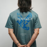 Person wearing a teal t-shirt with blue graphic of a dove bird and text Gwangbok in white on the back, standing against a plain background.
