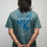 Person wearing a teal t-shirt with blue graphic of a dove bird and text Gwangbok in white on the back, standing against a plain background.