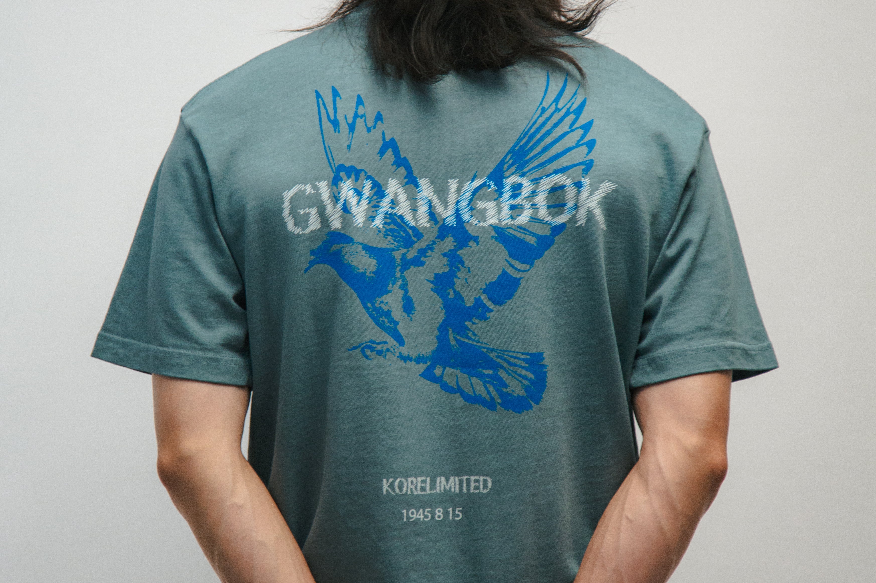 Person wearing a teal t-shirt with blue graphic of a dove bird and text Gwangbok in white on the back, standing against a plain background.