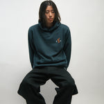 Person wearing a teal hoodie and black pants sitting on a white background