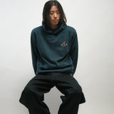 Person wearing a teal hoodie and black pants sitting on a white background