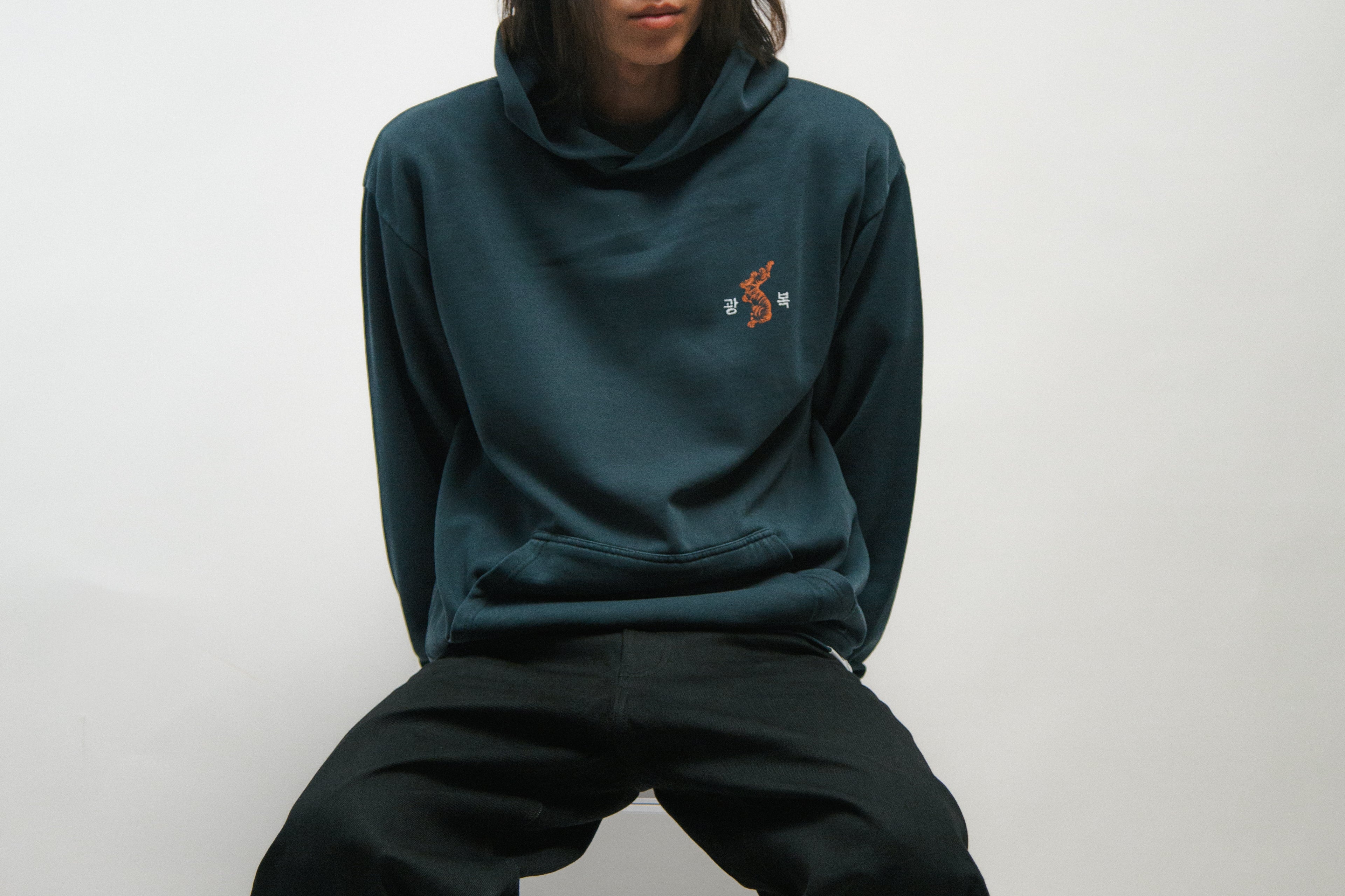 Person wearing a teal hoodie and black pants sitting on a white background