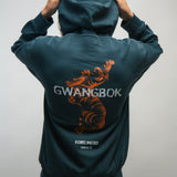 Person wearing a teal hoodie with a Korean tiger design and 'GWANGBOK' text on the back, against a plain background.