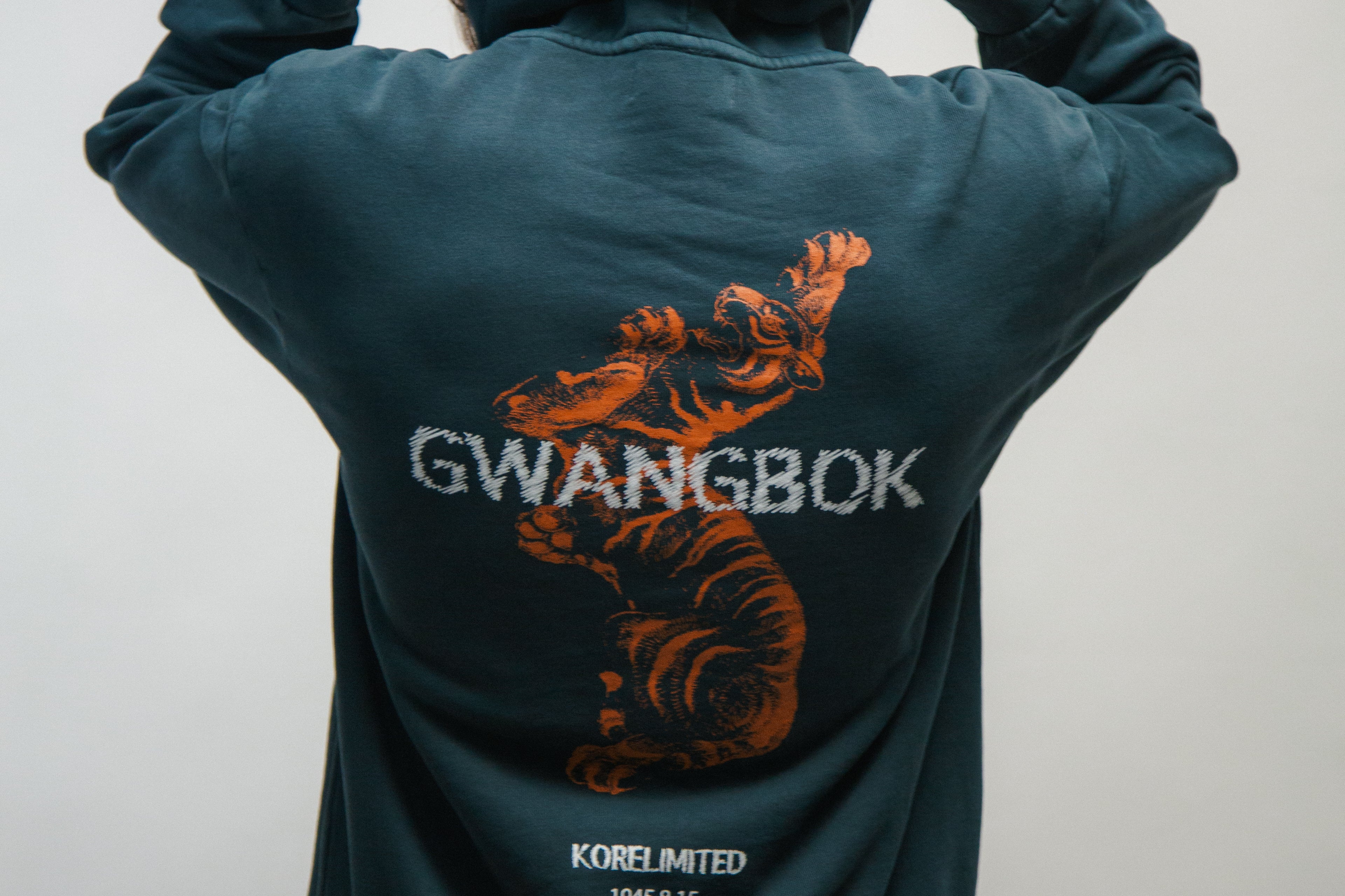 Person wearing a teal hoodie with a Korean tiger design and 'GWANGBOK' text on the back, against a plain background.