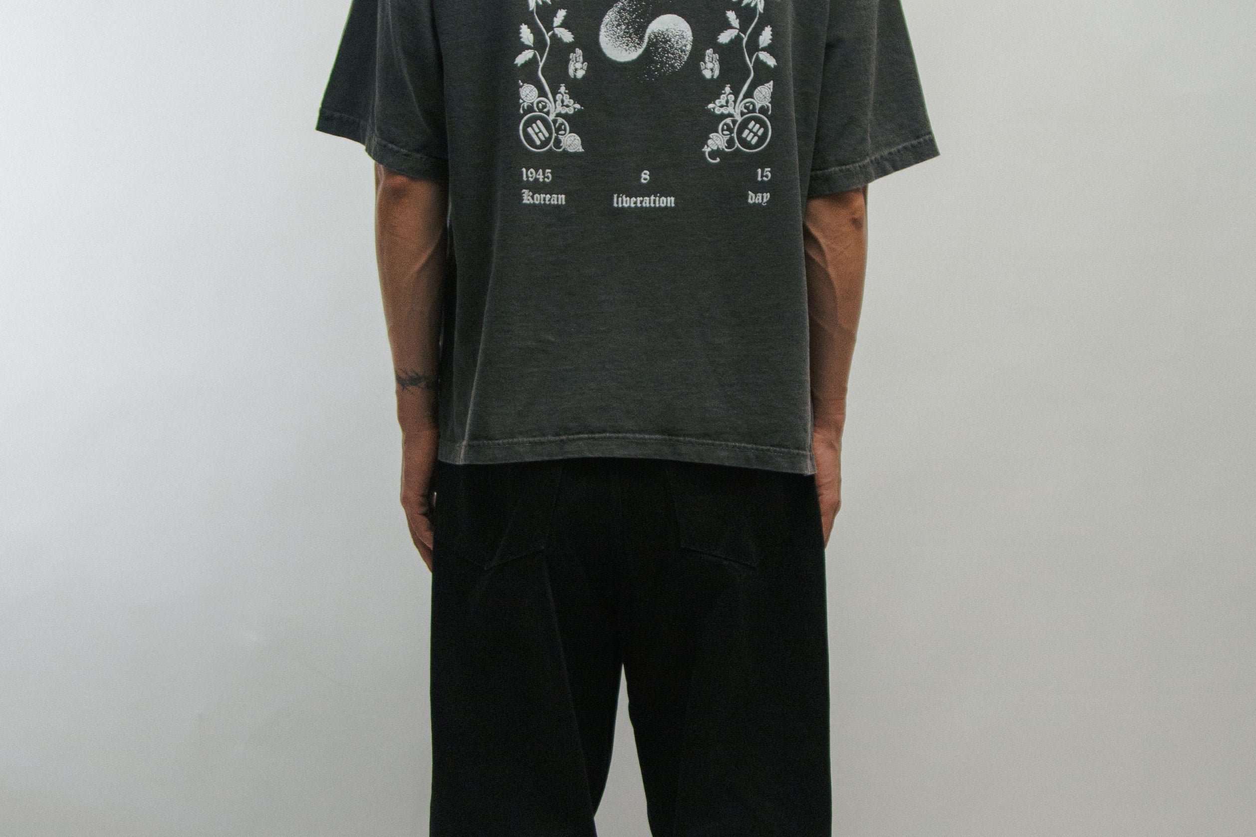 Person wearing a dark gray t-shirt with floral designs and text on the back for Korean Liberation Day, standing against a plain background.