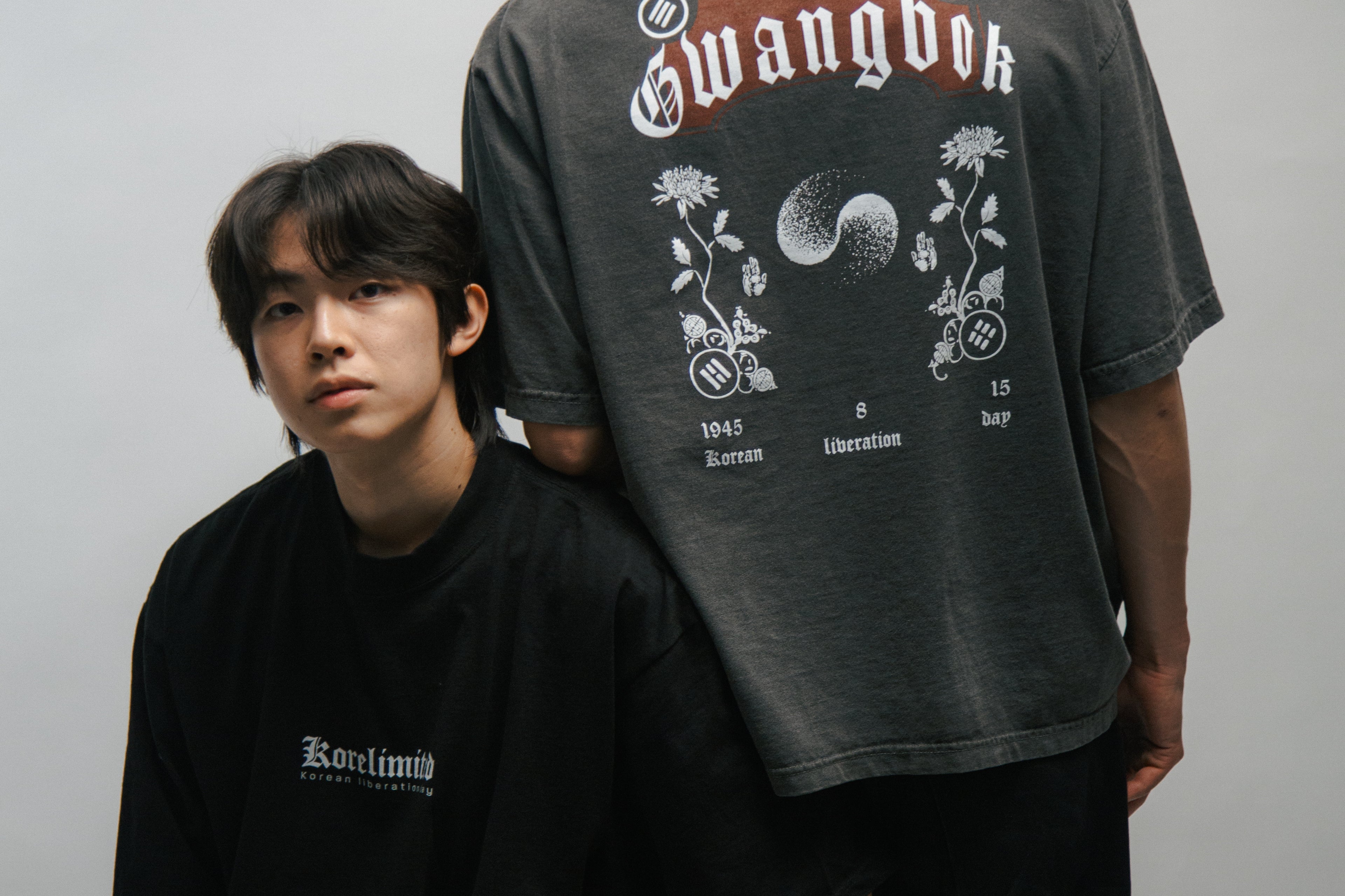 Two people wearing black and gray t-shirts with KORELIMITED and GWANGBOK graphic designs on a plain background