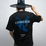 Person wearing a black t-shirt with blue dove graphics and white text Gwangbok, holding a large black Korean hat called gat.
