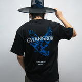 Person wearing a black t-shirt with blue dove graphics and white text Gwangbok, holding a large black Korean hat called gat.