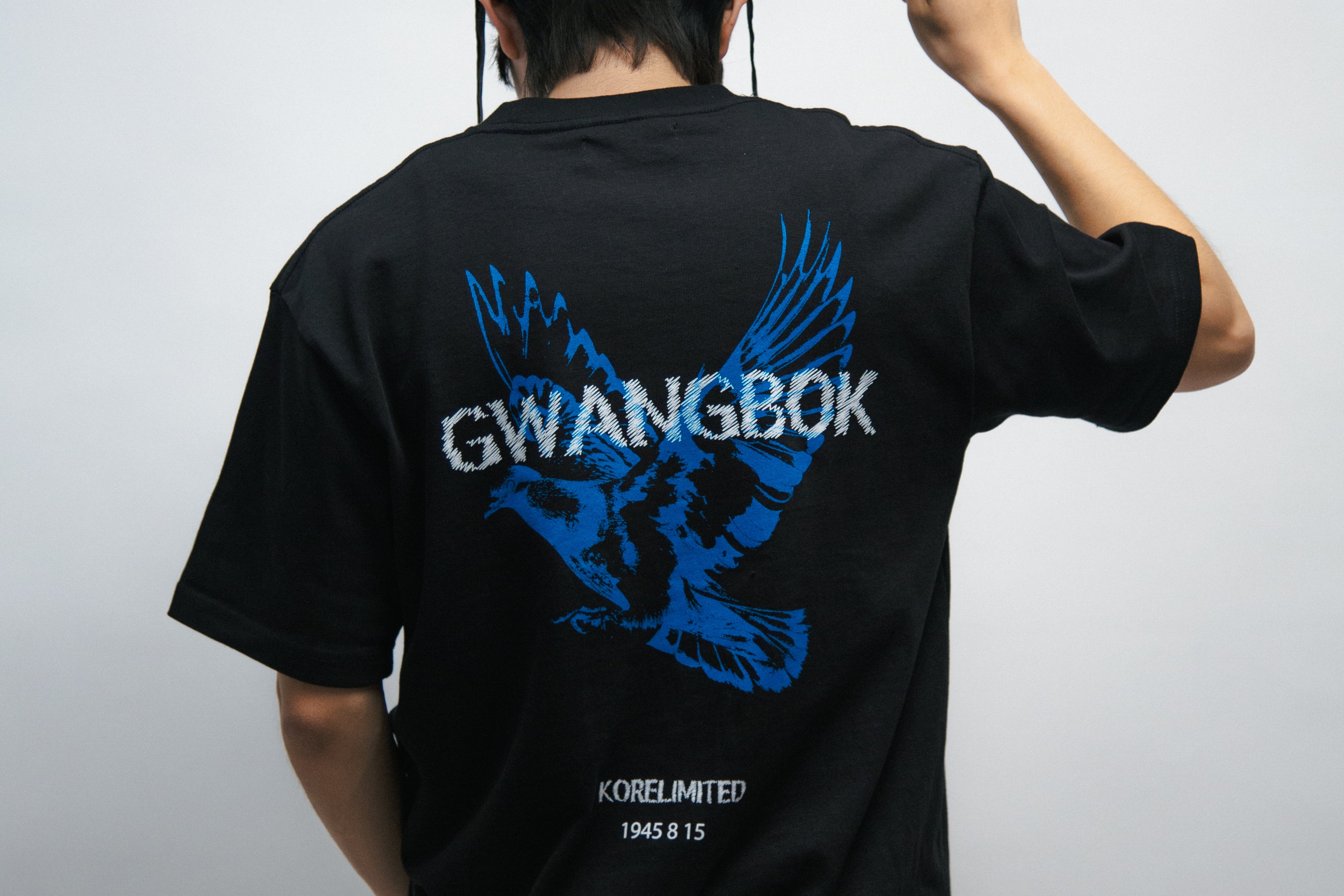 Person wearing a black t-shirt with blue dove graphics and white text Gwangbok, holding a large black Korean hat called gat.