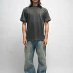 Male person wearing a dark gray t-shirt with Korean characters and blue jeans standing against a plain background