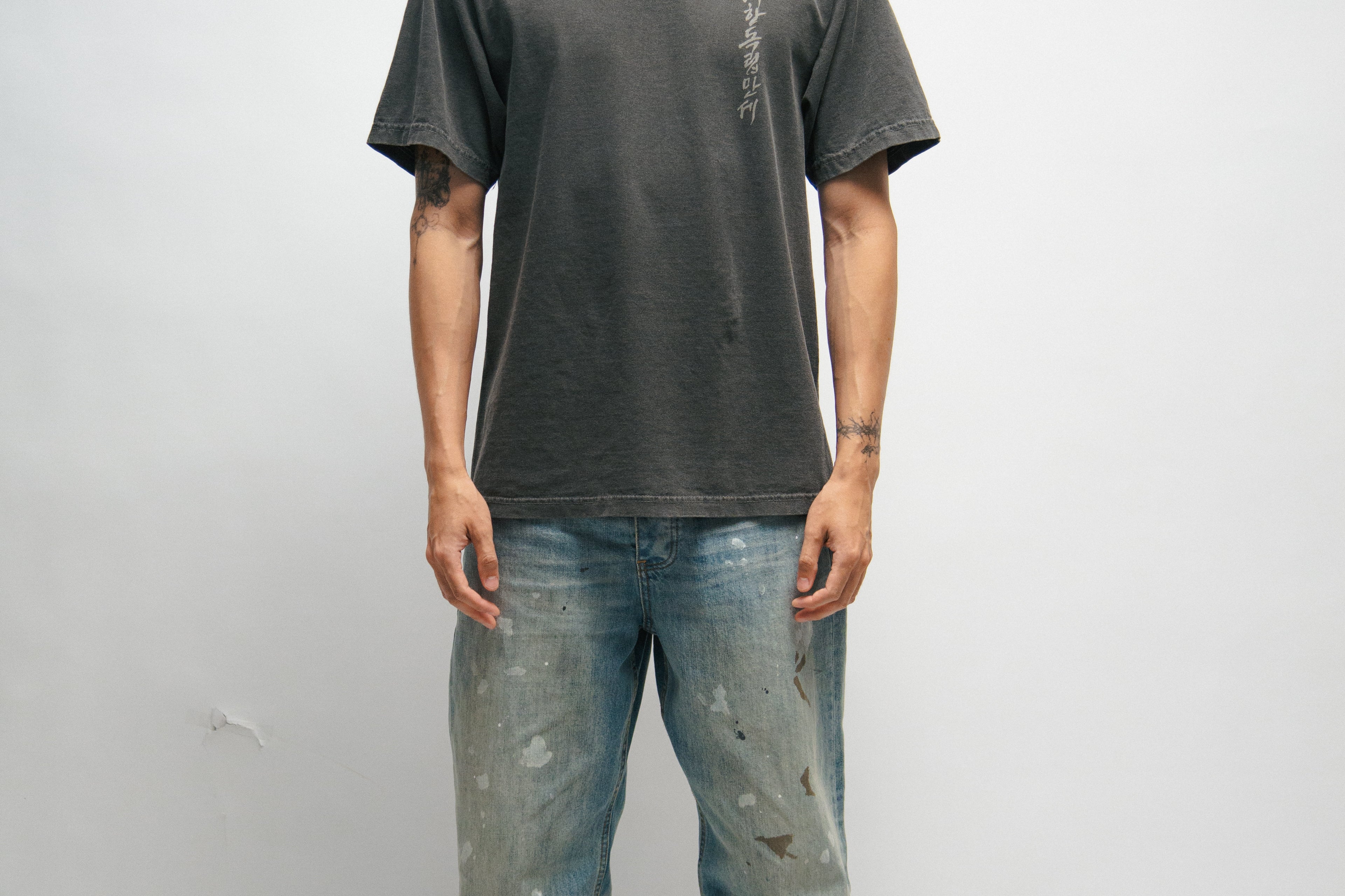 Male person wearing a dark gray t-shirt with Korean characters and blue jeans standing against a plain background