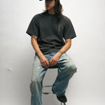 Person sitting on a stool wearing a dark grey t-shirt with Korean font, blue jeans, and a black cap against a white background