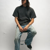 Person sitting on a stool wearing a dark grey t-shirt with Korean font, blue jeans, and a black cap against a white background