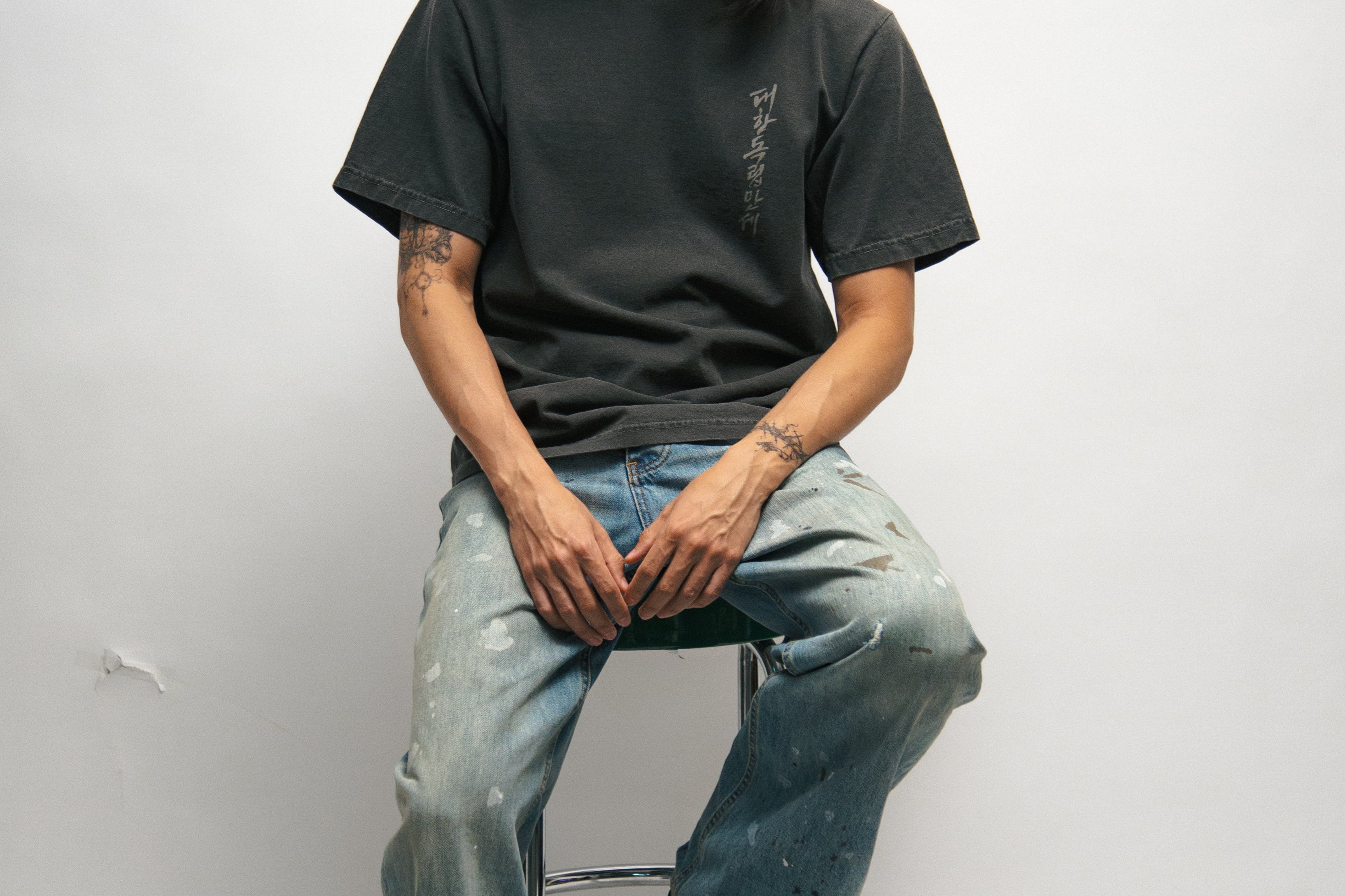 Person sitting on a stool wearing a dark grey t-shirt with Korean font, blue jeans, and a black cap against a white background