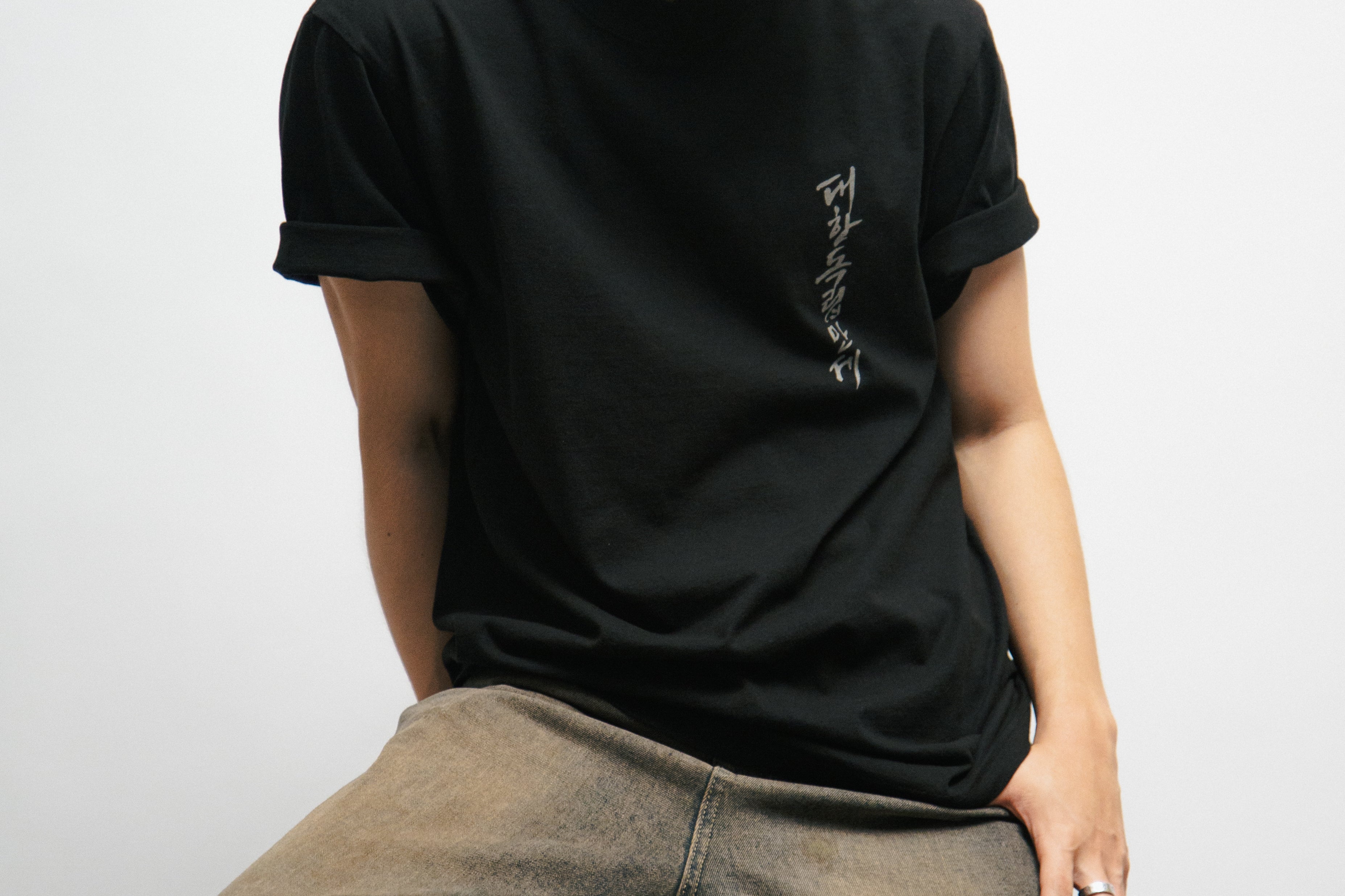 Person wearing a black t-shirt with Korean font on the front and cap with text, sitting against a white background